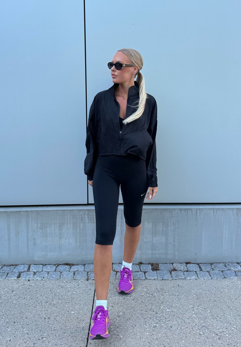 Femme avec queue de cheval blonde portant des lunettes de soleil noires, une veste noire, un short de cyclisme noir, des chaussettes blanches et des baskets violettes marchant dehors.