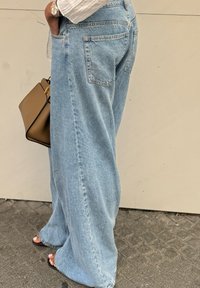 Jeans de pernera ancha en un azul claro, con una textura desgastada, que cuentan con bolsillos traseros, combinados con un bolso marrón y una blusa blanca sutil.