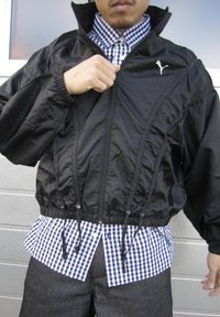 Mężczyzna zapinający czarną kurtkę Puma na niebiesko-białą koszulę w kratę i ciemne spodnie, stojący na tle jasnej ściany.