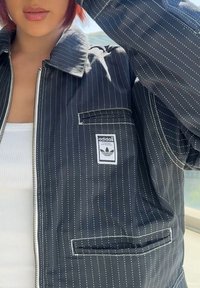 Chaqueta negra con cremallera y rayas blancas, con un bolsillo de parche y etiqueta del logo de Adidas en el pecho izquierdo. Textura suave y diseño con cuello.
