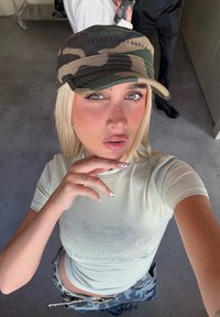 Beige fitted shirt with rhinestone text, camo cap with a curved brim, and patterned cargo pants. Polished nails with black and white accents.