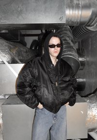 Giacca bomber nera con cappuccio, indossata sopra una maglietta nera. Jeans azzurri chiari, in piedi davanti a uno sfondo industriale metallico. Occhiali da sole scuri.