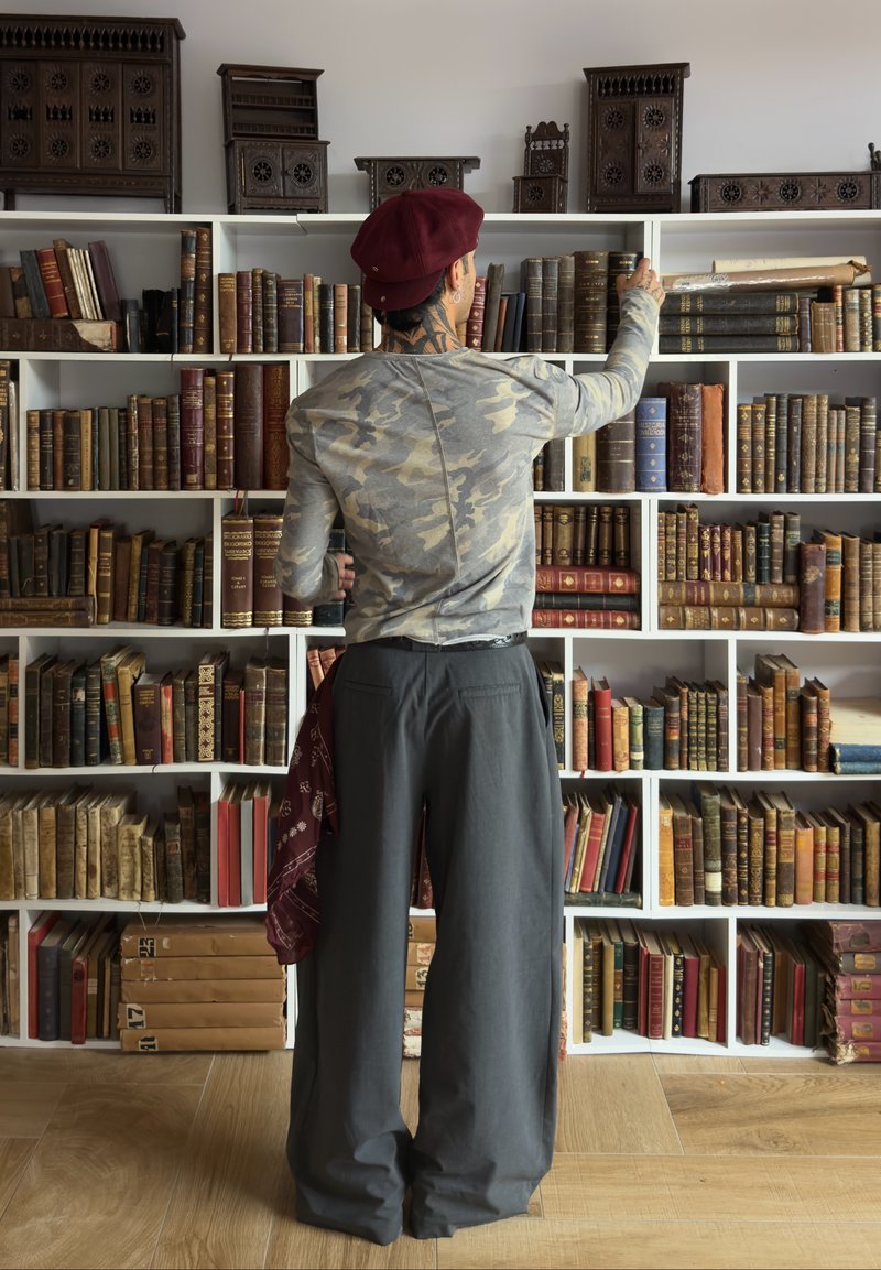 Borsa a tracolla, camicia a maniche lunghe grigia a motivo mimetico, pantaloni larghi scuri e un fazzoletto rosso. Librerie in legno piene di vari libri antichi.