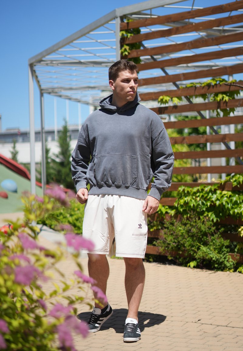Junger Mann in grauem Kapuzenpullover und weissen Shorts, der draußen bei einem hölzernen Rankgerüst mit grünen Reben unter klarem blauem Himmel spaziert.