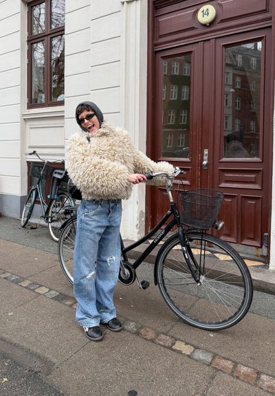 Persona con gafas de sol y una chaqueta beige esponjosa, de pie en una acera sosteniendo una bicicleta negra cerca de un edificio con una puerta roja con el número 14.
