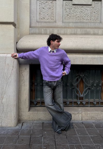 Hombre con suéter morado y vaqueros anchos negros apoyado contra una pared de piedra con una reja metálica decorativa en la ventana detrás de él.