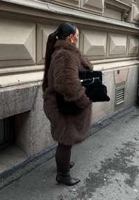 Brun fuskpälsjacka, ribbade bruna ärmar, svart handväska med silverdetaljer, bruna strumpbyxor och bruna klackstövlar, mot en texturerad vägg.