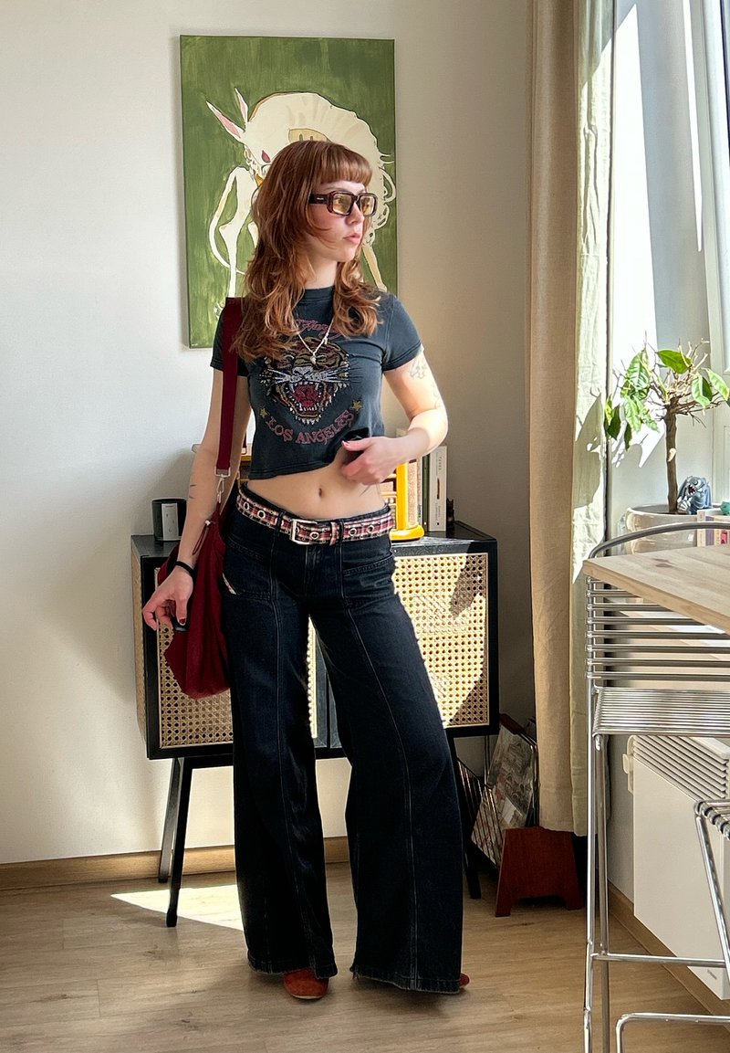 Jeune femme avec des lunettes de soleil, un crop top graphique tigre, un jean à jambes larges et un sac rouge posant à l'intérieur près d'une plante et d'une armoire.