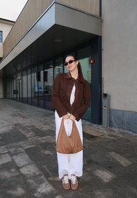 Jeune femme portant une veste marron et des lunettes de soleil tient un sac beige, debout sur un passage extérieur carrelé à côté d'un bâtiment moderne en verre.