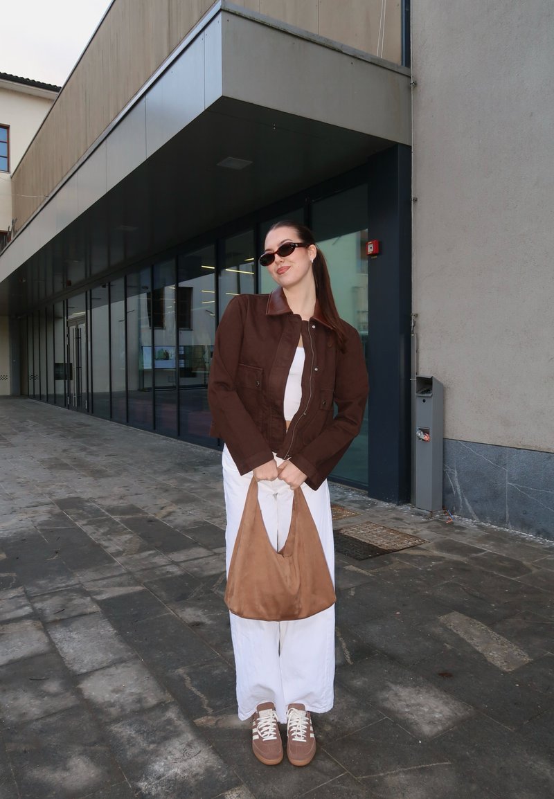 Jeune femme portant une veste marron et des lunettes de soleil tient un sac beige, debout sur un passage extérieur carrelé à côté d'un bâtiment moderne en verre.