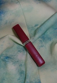 Tube de rouge à lèvres L'Oréal en rouge profond, de forme cylindrique avec une texture mate, bandeau en accent argenté, posé sur un tissu pastel bleu et vert.