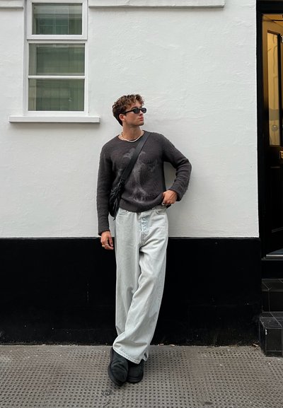 Suéter gris con un diseño texturizado, pantalones de mezclilla anchos de color claro, bolsa cruzada negra y calzado negro. El fondo de la pared es blanco con ribetes negros.