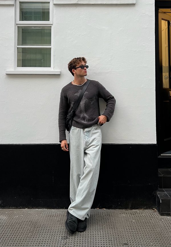 Sweater cinza com um design texturizado, calças largas de denim claro, bolsa preta tiracolo e calçado preto. O fundo da parede é branco com contornos pretos.