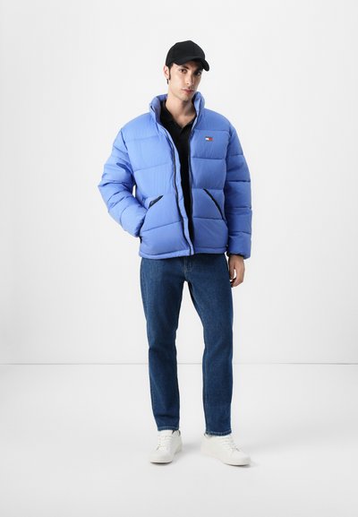Chaqueta acolchada azul claro con textura acolchada, acentos negros y un parche de logo. Lleva puesta sobre una camiseta negra con jeans azules y zapatillas blancas.