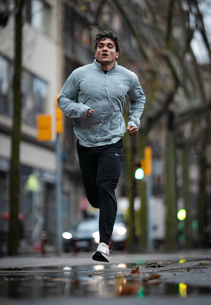 Άνδρας με γκρι μπουφάν και μαύρο παντελόνι τρέχει σε υγρό δρόμο της πόλης, με τα θαμπά κτίρια και τα φανάρια να φαίνονται στο παρασκήνιο.
