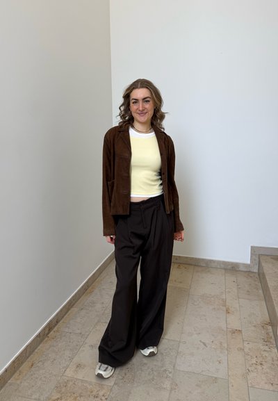 Mujer joven sonriendo, con chaqueta marrón, top amarillo pálido, pantalones anchos negros y zapatillas, de pie en una zona luminosa de escaleras interiores.