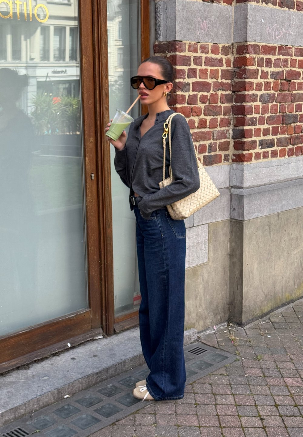 Maglietta scura a maniche lunghe, jeans blu a vita alta, borsa beige intrecciata con tracolla a catena e scarpe beige senza lacci. Tiene in mano una bevanda verde.