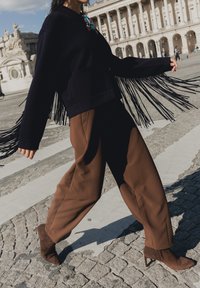 Personne marchant sur une rue pavée, portant un pantalon marron, des bottes marron à talons et une veste noire à franges avec une broche.