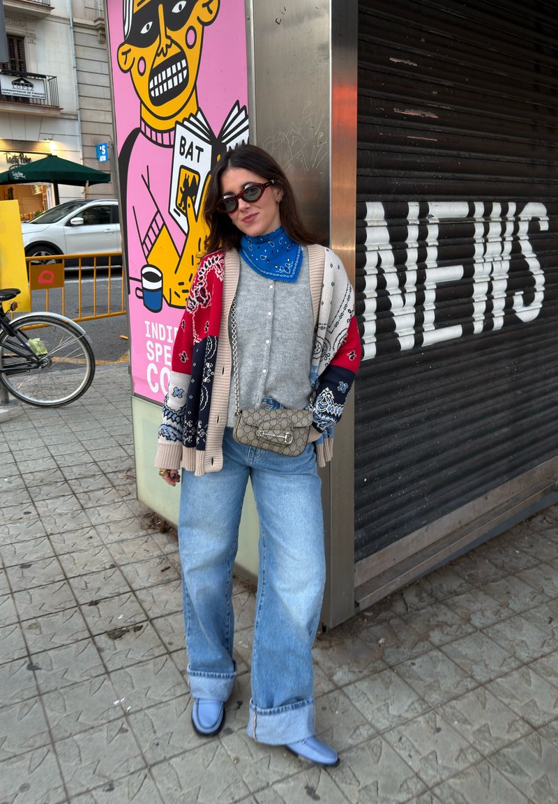 Femme portant des lunettes de soleil, un bandana bleu, un cardigan à motifs, une chemise grise, un jean large et des chaussures bleues, debout près d'un volet avec le texte "BARE PUB".