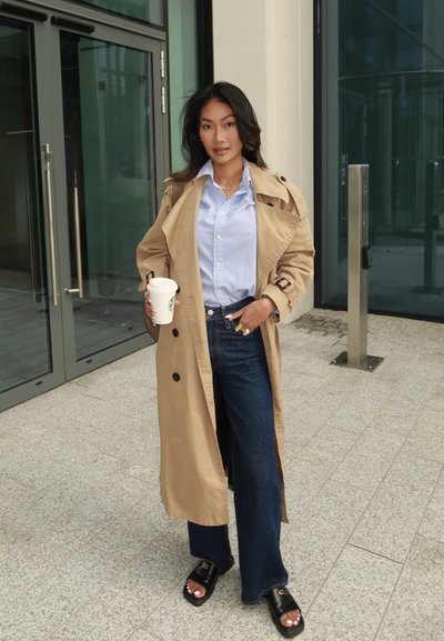 Un abrigo trench beige sobre una camisa de rayas azul, combinado con unos jeans oscuros de pierna acampanada y sandalias negras tipo slide. Sosteniendo una taza de café. Textura suave.