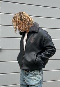 Must nahast pommitaja jope, millel on hall kunstkarusnahast krae ja vasakul rinnal embleem. Tõmblukk ja soonikkoes mansetid.