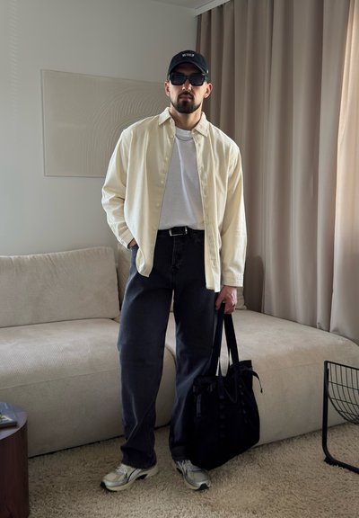 Hombre con gorra negra, gafas de sol, camisa beige sobre camiseta blanca, pantalones oscuros, zapatillas blancas, sujetando un bolso negro en una sala de estar.