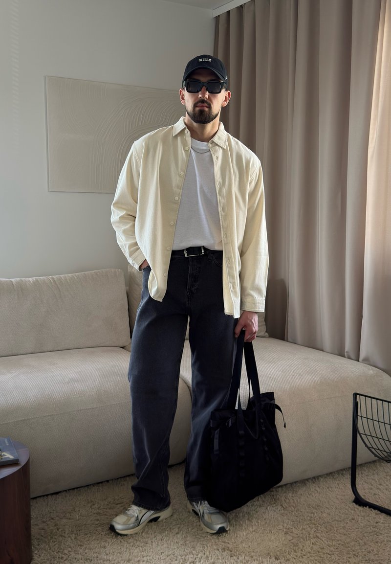 Homme portant une casquette noire, des lunettes de soleil, une chemise beige sur un t-shirt blanc, un pantalon foncé, des baskets blanches, tenant un sac fourre-tout noir dans un salon.