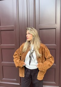 Brun semsket jakt med stor, komfortabel passform og trykknapper, kombinert med en hvit topp og et leopardmønstret skjerf. Svarte bukser fullfører antrekket.