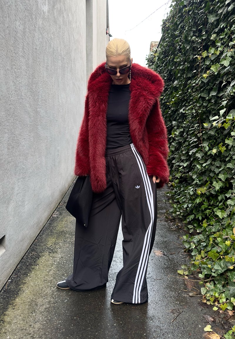 Personne portant un manteau en fausse fourrure rouge, chemise noire, pantalon large noir Adidas avec des bandes blanches, lunettes de soleil noires, tenant un sac noir, debout sur un chemin mouillé.