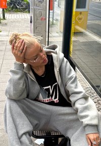 Grå joggingbukser og matchende hættetrøje lagdelt over en sort skjorte med hvidt logo. Dekorative ringe og tynde, prydede briller bæres.