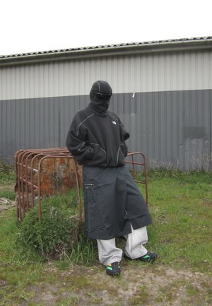 Persona que lleva pasamontañas negro, chaqueta de forro polar oscura, pantalones cargo anchos y deportivas, de pie al aire libre cerca de una puerta metálica oxidada y una pared de edificio.