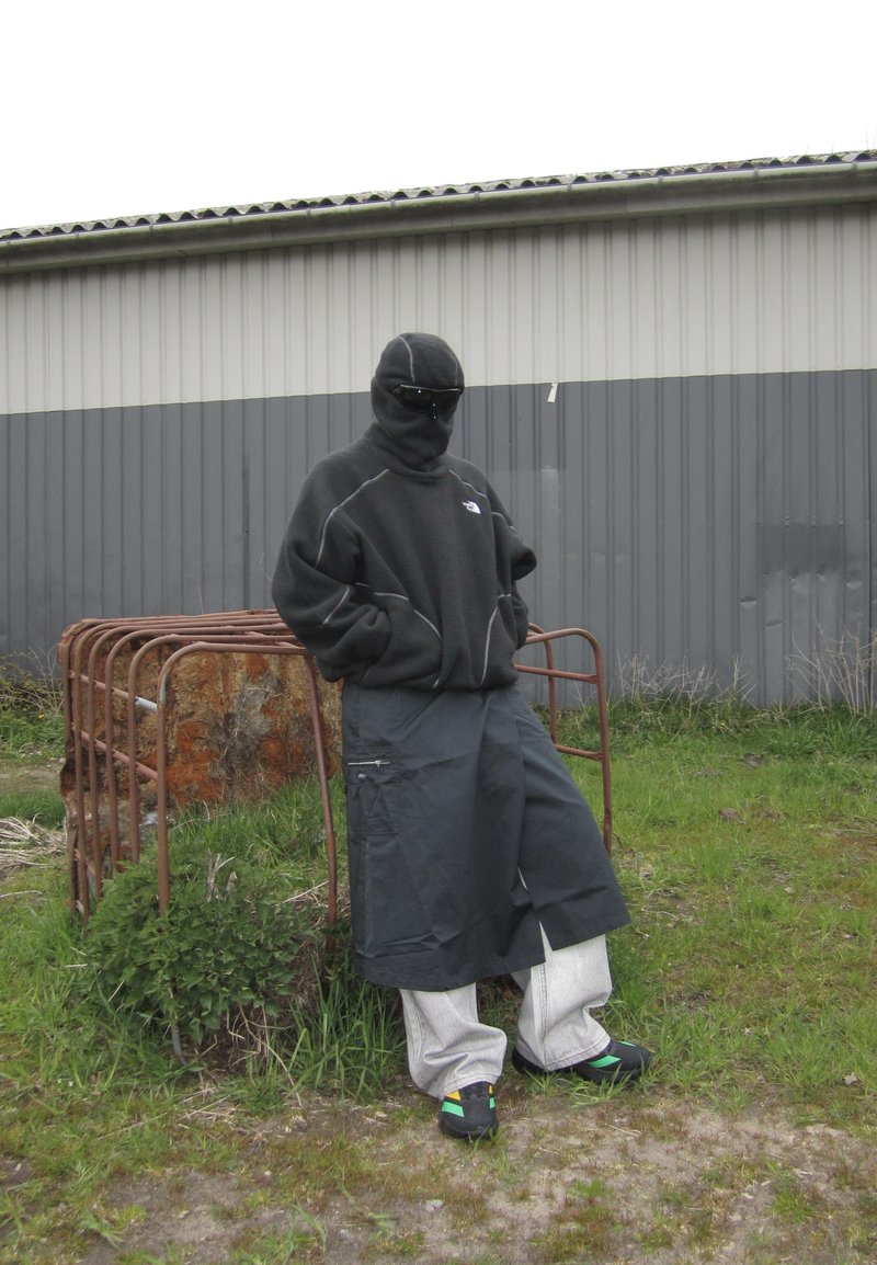 Personne portant un passe-montagne noir, une veste polaire foncée, un pantalon cargo large et des baskets, debout à l'extérieur près d'une porte métallique rouillée et d'un mur de bâtiment.