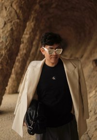 Svetlo béžový oversized blazer na čiernom tričku s logom, šedé nohavice a čierna kožená taška. Nosí biele slnečné okuliare s geometrickým rámom.