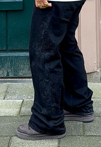 Černé kalhoty s květinovou výšivkou po straně, vyrobené z silné bavlněné tkaniny. Stylizované se šedými teniskami na kamenném chodníku.
