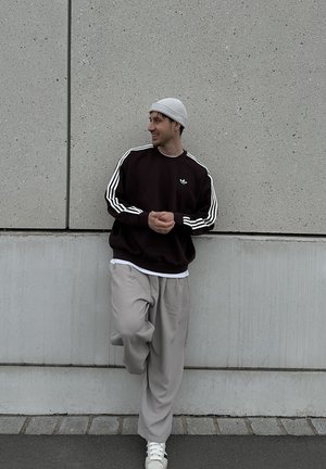 Hombre con gorro gris, sudadera negra con rayas blancas, pantalones beige y zapatillas blancas apoyado contra una pared de concreto, sonriendo y mirando de lado.