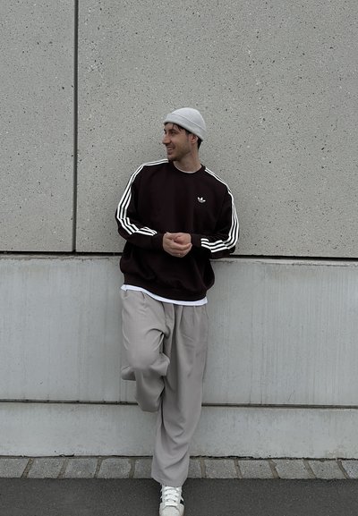 Hombre con gorro gris, sudadera negra con rayas blancas, pantalones beige y zapatillas blancas apoyado contra una pared de concreto, sonriendo y mirando de lado.