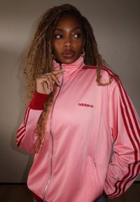 Jaqueta de treino adidas em satin rosa com detalhes vermelhos, gola alta, fecho de correr completo e três riscas em cada manga. Inclui bolsos laterais.