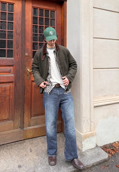 Hombre ajustándose una gorra verde, con chaqueta verde sobre una camisa a cuadros, suéter blanco, jeans azules y zapatos marrones, de pie junto a una puerta de madera y una pared de piedra clara.