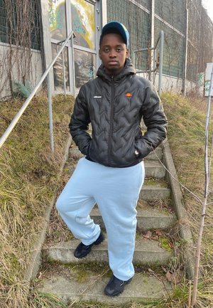 Chaqueta de plumón negra con un logo, pantalones de chándal azul claro y zapatillas negras. De pie en unos escalones de piedra con hierba y enredaderas de fondo.