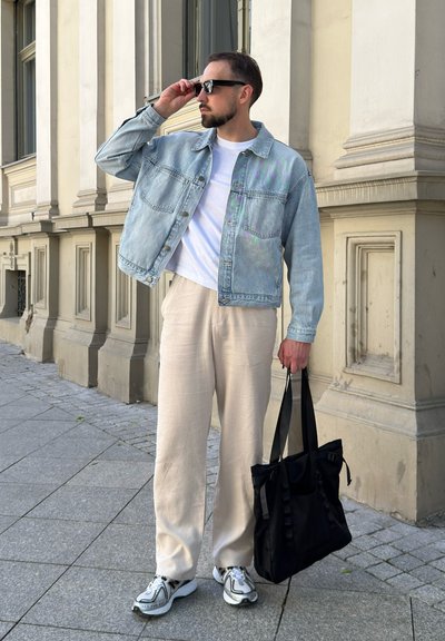 Hombre con gafas de sol, chaqueta vaquera clara, camiseta blanca, pantalones beige y zapatillas deportivas, sosteniendo un bolso tote negro en una acera de la ciudad.