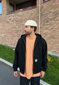 Black zip-up hoodie with large pockets, layered over a peach-coloured shirt featuring a Nike logo. The model also wears a light beige beanie.