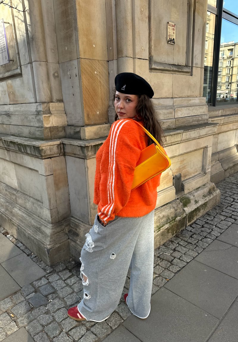 Femme portant un pull orange, un pantalon gris déchiré, un béret noir et un sac à main jaune, debout près d'un bâtiment en pierre dans une rue pavée.
