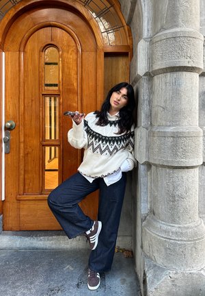 Gros pleten muzej s črno-belo geometrijsko vzorcem, modri široki kavbojki, rjavi športni copati, naslonjen ob teksturiranem kamnitm stebru.