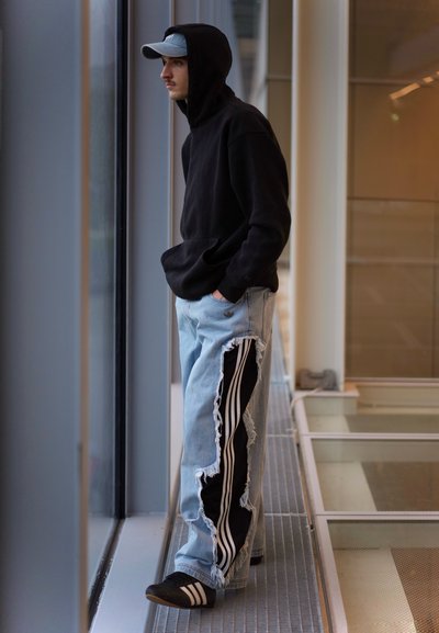 Joven con sudadera negra con capucha y vaqueros claros rotos con franjas negras laterales, de pie junto a la ventana mirando hacia fuera con las manos en los bolsillos.