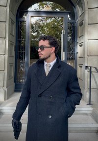 A man in a black overcoat, wearing sunglasses and a tie, stands with his hands in his pockets in front of a building entrance marked with the number 23.