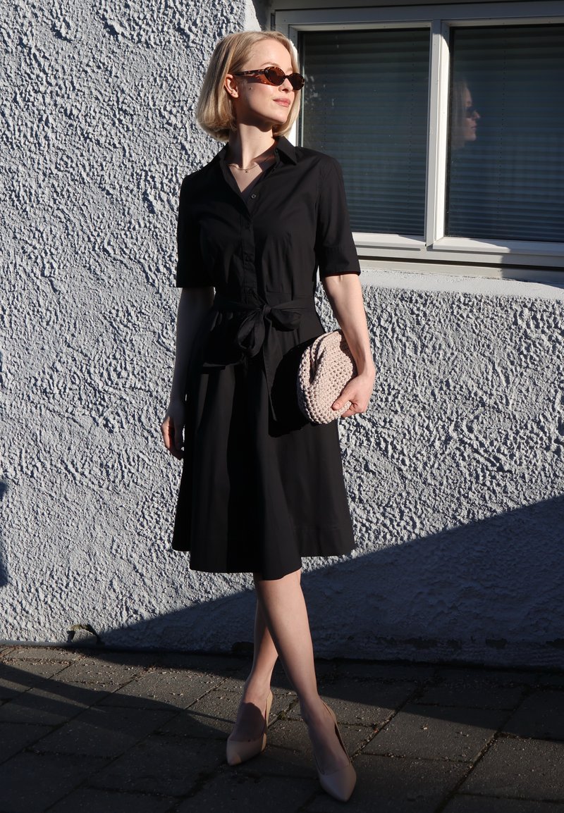 Femme portant une robe noire et des lunettes de soleil, tenant une pochette texturée, debout contre un mur blanc rugueux avec une fenêtre reflétant son image.