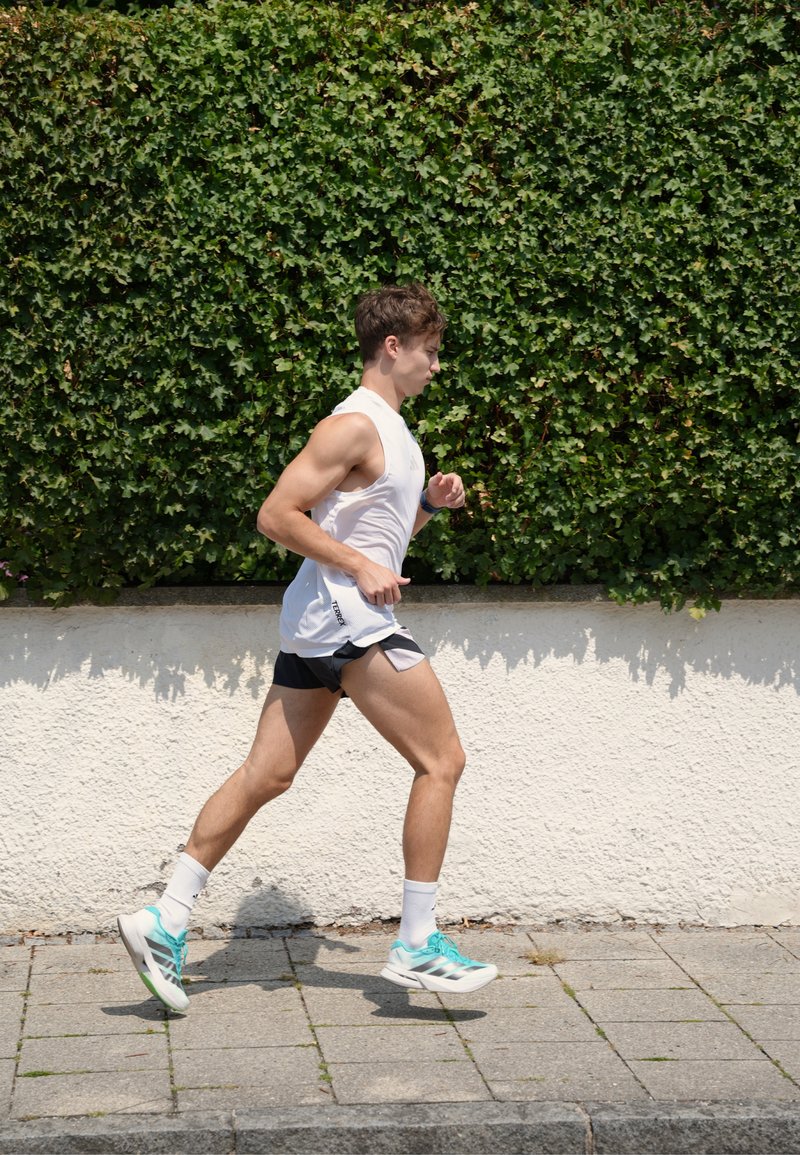 Giovane uomo che fa jogging su un marciapiede accanto a una siepe verde e frondosa, indossa una canottiera bianca, pantaloncini neri e scarpe da corsa azzurro chiaro.