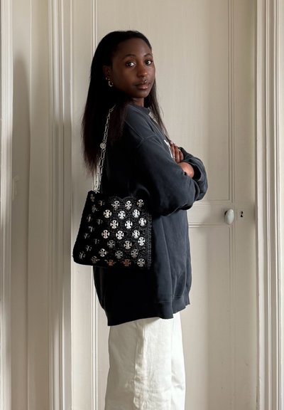 Mujer con cabello largo y oscuro que lleva una sudadera negra y pantalones blancos, con un bolso negro con estampado colgado del hombro, de pie en interiores.