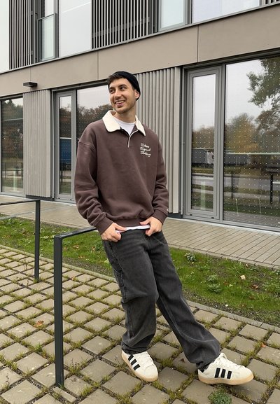 Sudadera marrón con cuello blanco, pantalones cargo grises y zapatillas blancas con rayas negras. De pie al aire libre cerca de un edificio moderno.