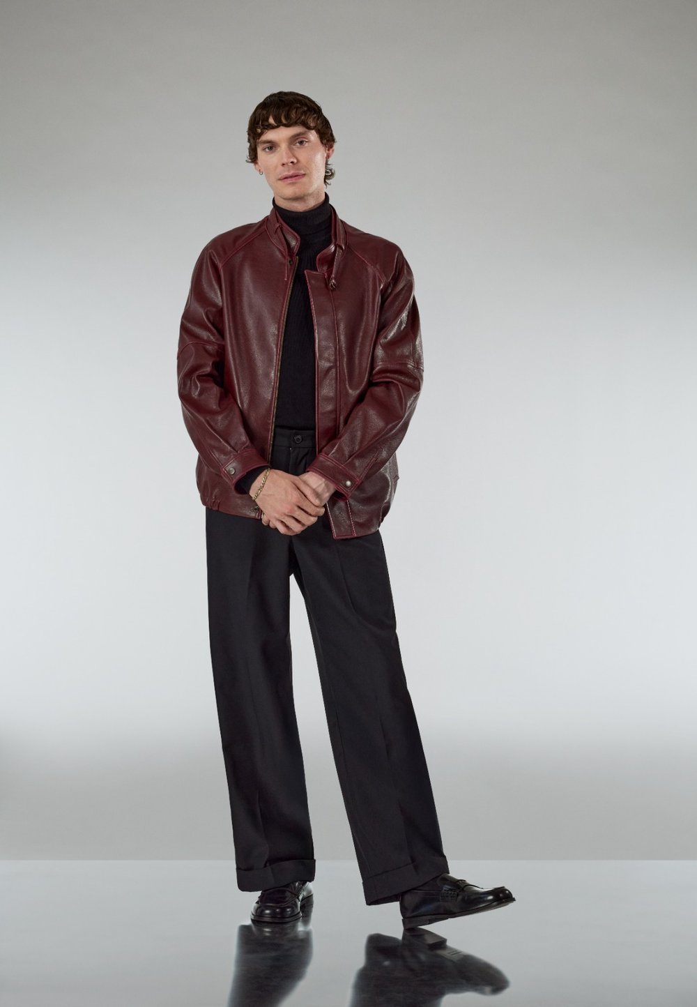 Burgundy leather jacket with zip fastening and stand collar, teamed with a black turtleneck, black trousers, and polished black shoes.